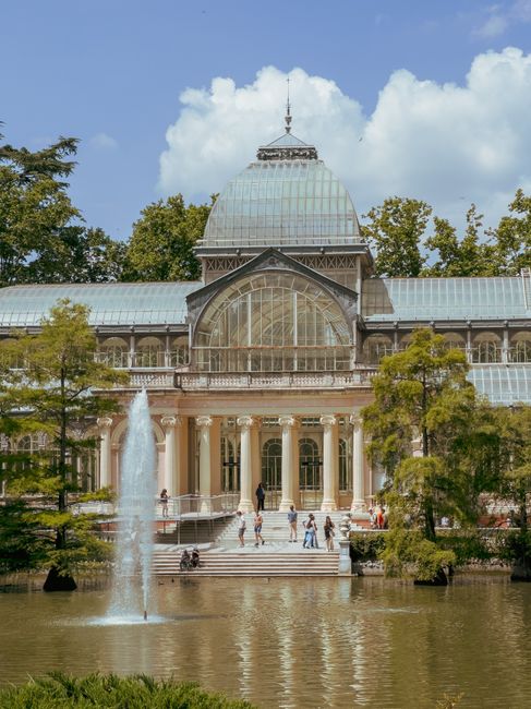 A large, ornate glass and iron greenhouse building located in Crystal Palace Park, with a central fountain in the pond in front of it. The structure features a curved glass roof with a decorative spire at the top, and multiple arched windows along its facade. Surrounding the building are tall trees with green foliage, some partially obscuring the structure. The scene is set on a clear day with blue skies and scattered white clouds. In the vicinity, visitors can be seen walking on the paved area leading up to the entrance, and some people are seated near the pond. For home relocation or furniture transport purposes, the image highlights the park's accessible pathways and open spaces that can facilitate loading or unloading items during removals, while the historic greenhouse provides a scenic backdrop for planning moving logistics near Crystal Palace. Removal Van Crystal Palace occasionally supports clients with such relocation needs within park surroundings.
