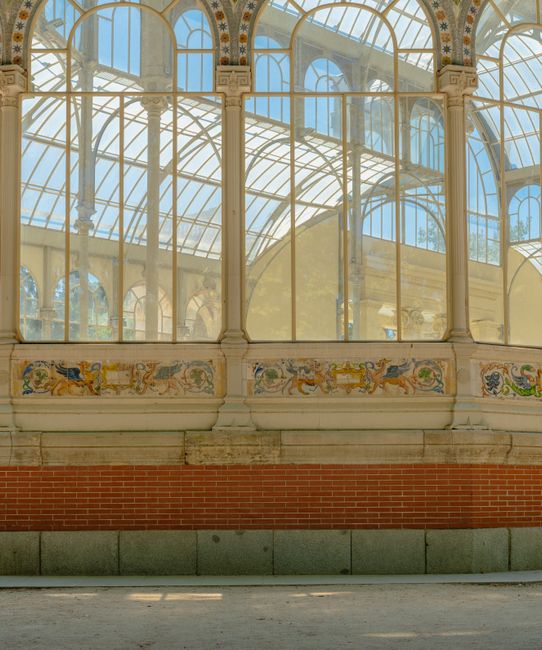 Detailed view of an ornate glass conservatory or greenhouse interior with large arched windows and decorative painted panels along the lower wall. The structure features a metal framework supporting expansive glass panes, allowing natural light to fill the space. The base of the building consists of a brick and stone foundation. This setting may be part of a historic or botanical property where furniture and boxes could be loaded or unloaded during home relocation or moving services. The image captures the architectural details and spacious environment suitable for furniture transport and packing activities involved in professional removals, as seen in services provided by Removal Van Crystal Palace, especially in the context of house removals and move logistics.