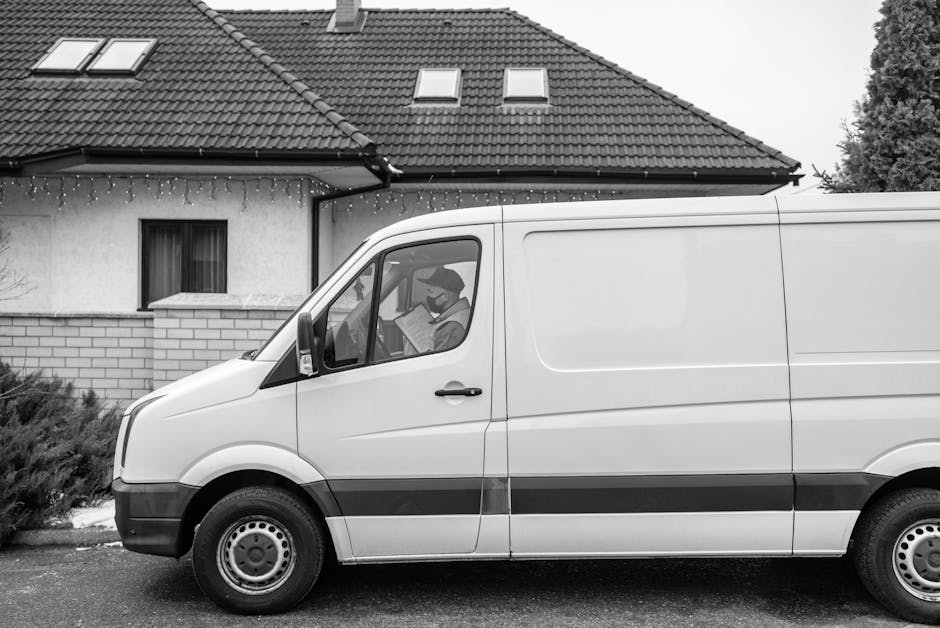 A man wearing a black and grey cap, black jacket, and dark trousers is seen carrying a medium-sized cardboard box with a label on top, during a home relocation process. He is outside a modern industri