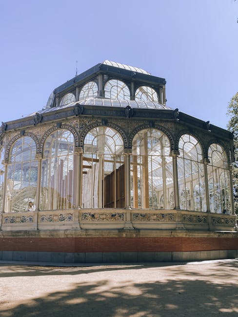 The image shows a close-up view of a historic glass and metal conservatory or greenhouse structure with ornate arched windows and intricate detailing along the exterior. The building features a large,