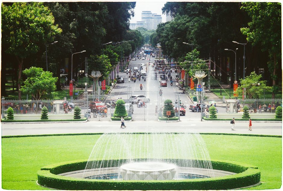 A park scene featuring a large circular fountain with multiple streams of water arching upwards in the foreground, surrounded by neatly trimmed grass and low hedges. Beyond the fountain, there is a wi